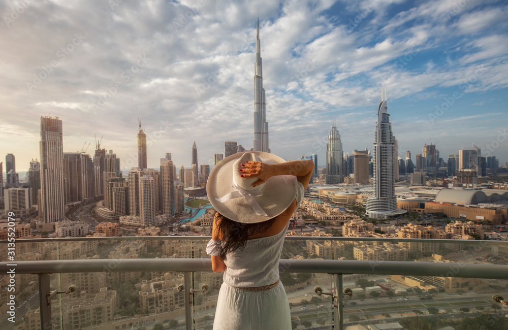 Woman with a white hat is standing on a balcony in front of the skyline from Dubai Downtown - obrazy, fototapety, plakaty 