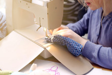 Enthusiastic Young Girl Works Concentrated With A Compact Sewing Machine Under Supervision Of Her Mother To Sew A Toy Dress For Her Doll - Background Blanked Out Blurry