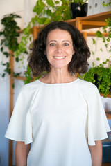 Cheerful middle aged Caucasian woman looking at camera. Front view of beautiful mature woman wearing white blouse standing at office. Female beauty concept