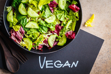 Wall Mural - Vegan food concept. Green vegetable salad in black plate and notebook with the word vegan, top view.