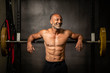 © Paul - Muscular shirtless mature older bodybuilding athlete with balding gray hair  leaning against a barbell in a gym smiling at the camera
