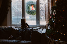 A Little Girl Sits Next To A Christmas Tree And Looks Out A Window.