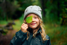 Long Haired Charming Girl In Warm Hat Closing Eye With Green Leaf In Forest