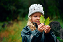 Charming Girl In Warm Hat Holding Green Leaves In Forest
