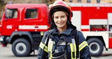 Portrait Of Young Woman Firefighter Standing Near Fire Truck. Fireman In Protective Suit With Oxygen Mask And Helmet.