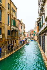  Venice, Italy. Rainy day in the city. Narrow water canal between gothic buildings and a bridge connects islands. People/ Tourists walk with umbrellas. Boats docked and sailing.