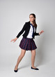 full length portrait of a pretty brunette girl wearing a school uniform of black jacket and plaid skirt. Standing pose isolated against a studio background.
