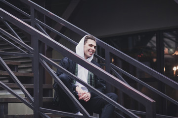 Wall Mural - Handsome young man hipster in jacket with hood sitting on a staircase