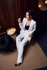 Wall Mural - Beautiful young pretty woman bright evening makeup of shiny red lipstick brunette curly hair wear elegant suit white trousers blouse jacket high heels clothes for party meeting interior room fashion.