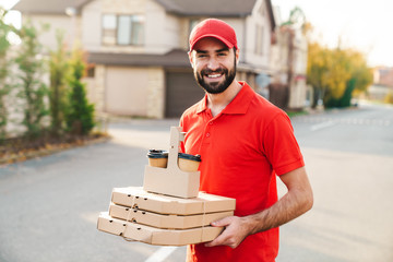 Wall Mural - Image of joyful young delivery man holding pizza boxes and coffee