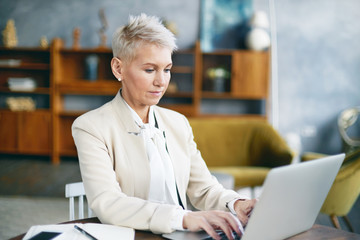 People, age, electronic gadgets and modern lifestyle concept. Confident successful mature female with dyed pixie hairstyle using wireless internet on laptop, checking email, typing business letter