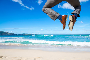 Businessman jumping into the air above a tropical beach clicking his heels together in celebration