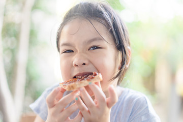 Wall Mural - Funny little asian girl eating tasty toasts with strawberry jam spread for breakfast. Hungry face and Delicious face in kid girl.