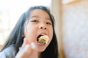 Wall Mural - Hungry face and enjoy eating concept.Little asian girl enjoy eating with cheese cake with fork on a plate in breakfast time at home.Concept for food, Dental care, Oral health.