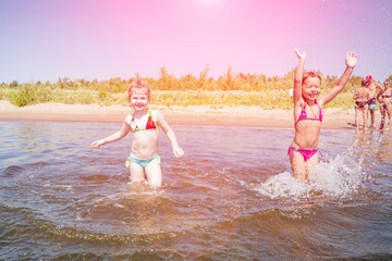 Wall Mural - Little girl friends run along the river and spray water on a sunny summer day.