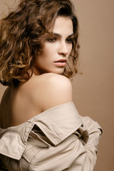 beauty fashion model with clean skin and curly hair in biege cloak stretch with scarf on biege background, bare shoulders