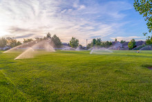 Sprinklers Watering Green Grassy Field With Homes And Cloudy Blue Sky Background