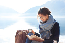 Young Woman Reading Book At Sea Beach. Cozy Winter Picnic By Morning Mountains. Happy Student In Blue Scarf Enjoying Traveling. Girl Traveler Drinks Coffee. Solo Female Tourism. Lifestyle Moment.