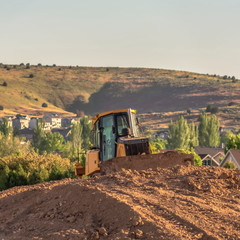 Wall Mural - Square frame Bulldozer on a mound of soil at a construction site against mountain and sky