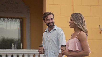 Poster - Side view of young positive couple man and woman are walking by street outdoors in summer holding a paper cups of tea or coffee