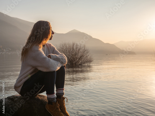 Fotografía Young woman witting on rock by the lake at sunset looking at view and enjoying the serene atmosphere