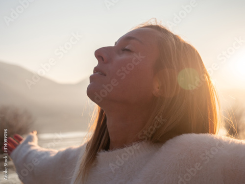 Happiness concept, woman by the lake at sunset enjoying golden hour breathing and inhaling fresh air from mountains. Young woman closing eyes, spirituality day dreaming, hope and faith