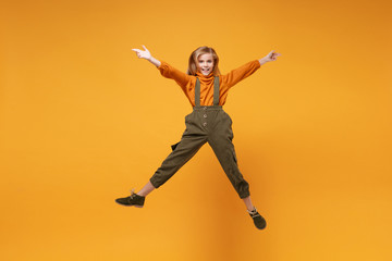 Smiling little kid girl 12-13 years old in turtleneck, jumpsuit isolated on orange yellow wall background in studio. Childhood lifestyle concept. Mock up copy space. Jumping, spreading hands and legs.