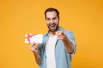 Poster - Laughing young man in casual blue shirt posing isolated on yellow orange background, studio portrait. People lifestyle concept. Mock up copy space. Hold gift certificate, point index finger on camera.