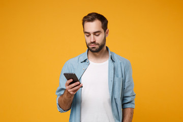 Young man in casual blue shirt posing isolated on yellow orange wall background, studio portrait. People sincere emotions lifestyle concept. Mock up copy space. Using mobile phone, typing sms message.