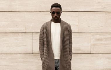 Wall Mural - Stylish african man wearing brown knitted cardigan, sunglasses, male model posing on city street over brick wall background