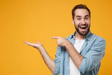 Poster - Excited young bearded man in casual blue shirt posing isolated on yellow orange background, studio portrait. People emotions lifestyle concept. Mock up copy space. Pointing index finger, hand aside.