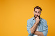 Pensive young bearded man in casual blue shirt posing isolated on yellow orange background studio portrait. People emotions lifestyle concept. Mock up copy space. Put hand prop up on chin, looking up.