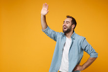 Cheerful Young Bearded Man In Casual Blue Shirt Posing Isolated On Yellow Orange Background In Studio. People Lifestyle Concept. Mock Up Copy Space. Waving And Greeting With Hand As Notices Someone.