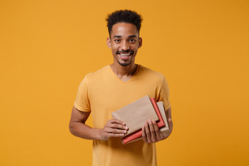 Joyful young african american guy in casual t-shirt posing isolated on yellow orange wall background, studio portrait. People lifestyle concept. Mock up copy space. Hold in hands books, notebooks.