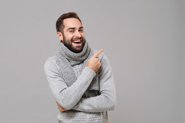 Poster - Laughing young man in gray sweater, scarf posing isolated on grey wall background, studio portrait. Healthy fashion lifestyle, cold season concept. Mock up copy space. Pointing index finger up aside.