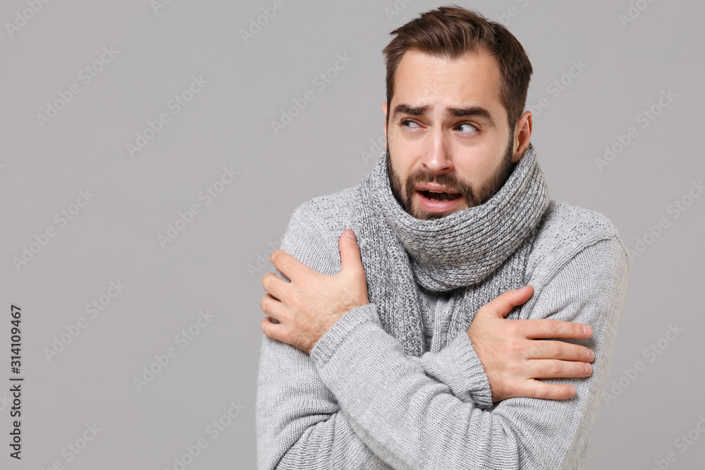 Frozen young man in gray sweater posing isolated on grey wall background, studio portrait. Healthy fashion lifestyle, cold season concept. Mock up copy space. Holding hands folded, huging himself. - obrazy, fototapety, plakaty 