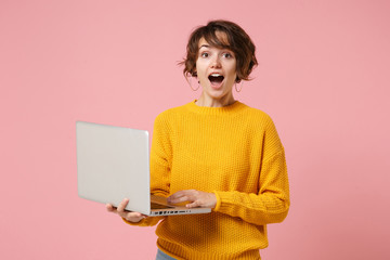 Shocked young brunette woman girl in yellow sweater posing isolated on pastel pink background studio portrait. People lifestyle concept. Mock up copy space. Holding and working on laptop pc computer.