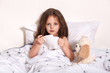 © sementsova321 - Portrait of exhausted ill infant looking directly at camera, holding handkerchief in both hands, lying in soft cosy bed with her teddy bear, suffering from running nose. Children and health concept.