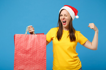 Wall Mural - Funny Santa girl in Christmas hat posing isolated on blue background. New Year 2020 celebration holiday concept. Mock up copy space. Hold package bag purchases after shopping, showing biceps muscles.