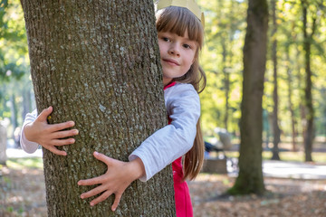 Portrait of a pretty smiling child girl in a paper crown on her head embracing a tree trunk in summer park. Love and care about nature concept.