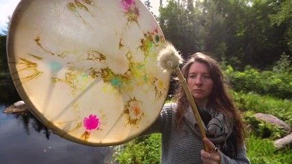 Wall Mural - Closeup and front footage of a woman with nose piercing and brunette hair playing a spiritual drum and beater. under bright summer sun in nature