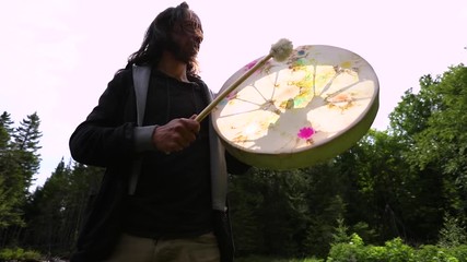 Wall Mural - Low angle and slow motion footage of a bohemian man playing a sacred native drum beneath bright summer sun. with lens flare and trees in background