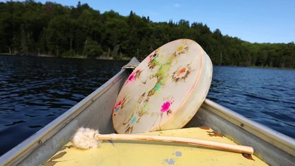 Wall Mural - Close up footage of a sacred native drum with dyed stretched membrane and traditional soft wood beater, floating in a canoe on a natural lake, with copy space