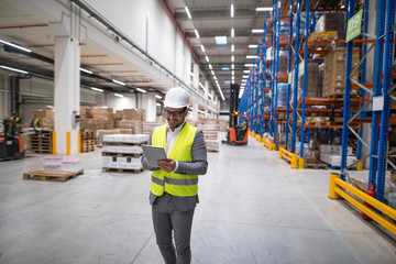 Wall Mural - Warehouse manager walking through large storage area and holding tablet while forklift operating in background.