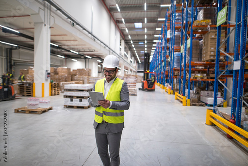 Cuadros en Lienzo Warehouse manager walking through large storage area and holding tablet while forklift operating in background