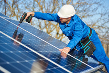 Man Worker In Blue Suit And Protective Helmet Installing Solar Photovoltaic Panel System Using Screwdriver. Electrician Mounting Module On Roof Of Modern House. Alternative Energy Ecological Concept.