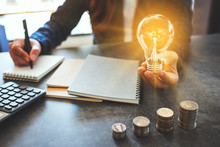 Businesswoman Holding A Lightbulb While Taking Note On Notebook With Coins Stack On Table, Saving Energy And Money Concept