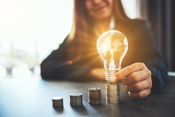 Wall Mural - businesswoman holding and putting lightbulb on coins stack on table for saving energy and money conc