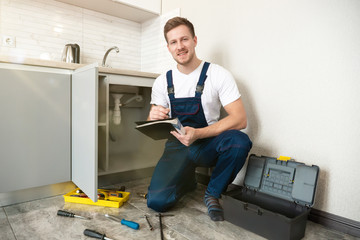 young handsome man plumber looks happy while taking notes in checklist during pipe fixing work in the kitchen professional plumbing repair service