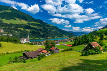 Wall Mural - Catholic church, traditional wood and modern houses along the lake Lungernsee in swiss village Lungern in sunny summer day, Obwalden, Switzerland